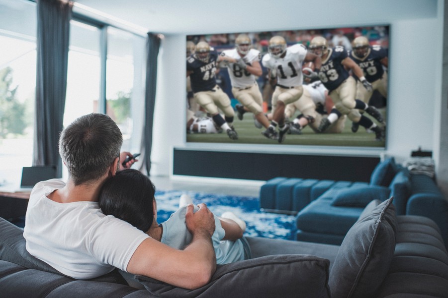 two people sit on a sofa watching football on a large screen TV mounted on the wall in front of them.